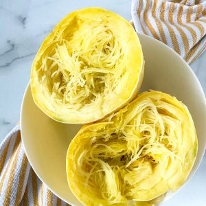 a cooked spaghetti squash cut in half in a white bowl on a yellow and white striped napkin