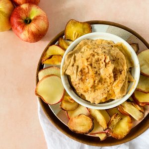 A bowl with pumpkin dip served with apple chips on the side with a pink background