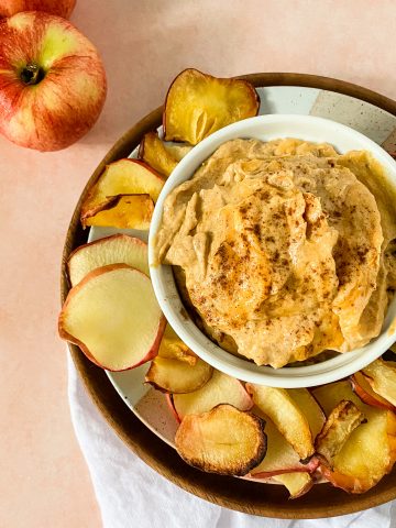 A bowl with pumpkin dip served with apple chips on the side with a pink background