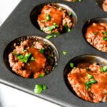 Square close-up of mini BBQ meatloaf cups in a dark muffin tin, topped with BBQ sauce and fresh herb garnish