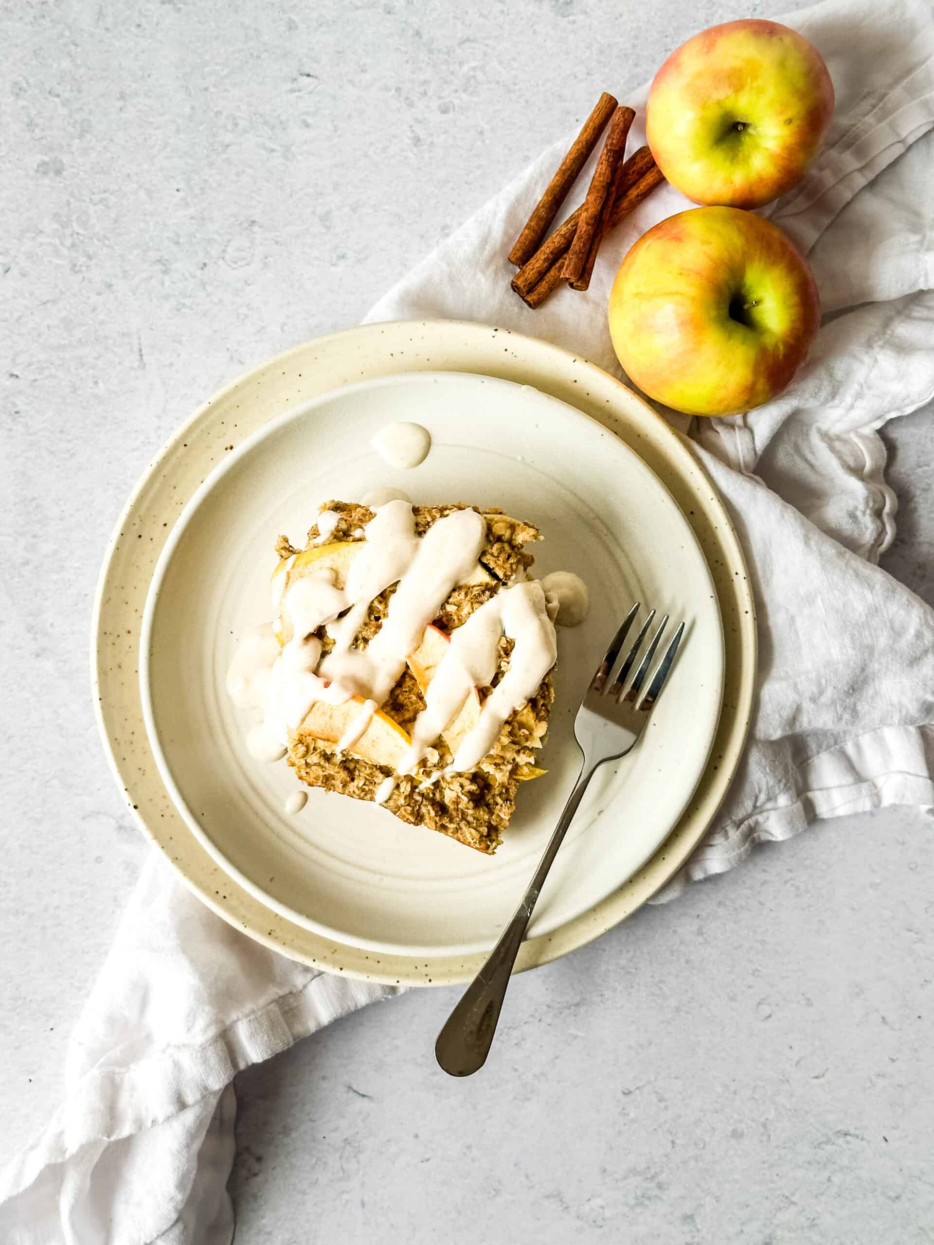 Slice of apple spice baked oatmeal with white glaze on a ceramic plate with a fork, whole apples, and cinnamon sticks in the background